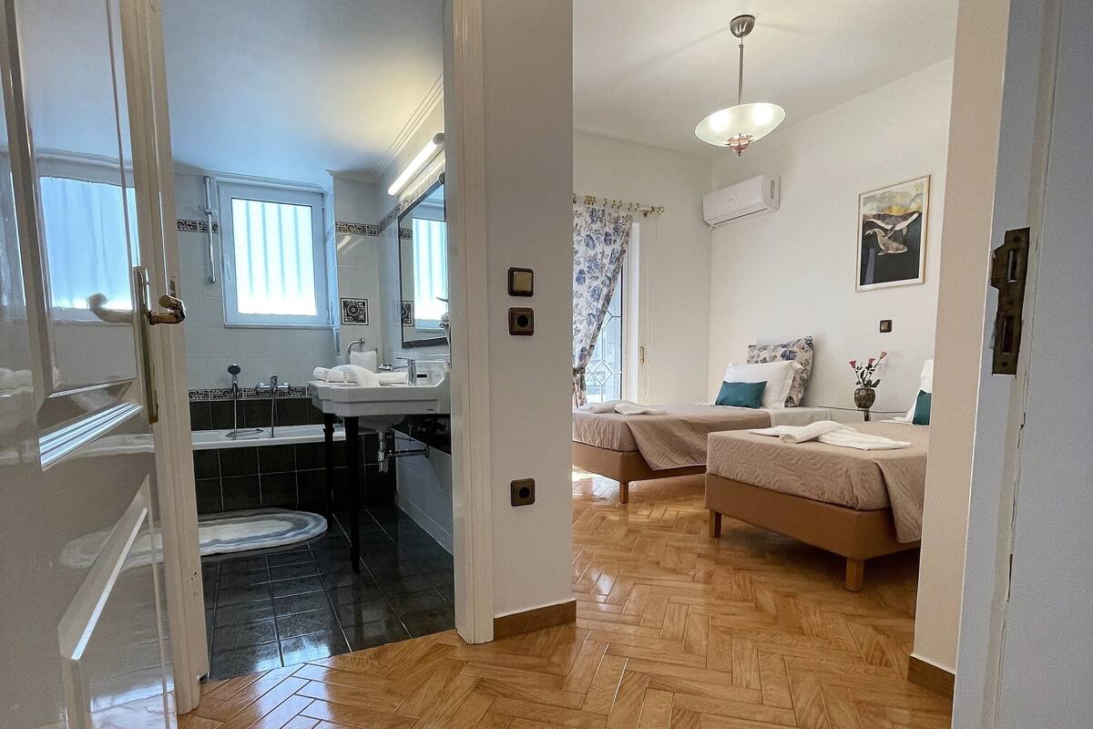 A hotel room with a wooden herringbone floor, a bed with a beige bedspread, a bedside table with a vase, and a window with curtains.
