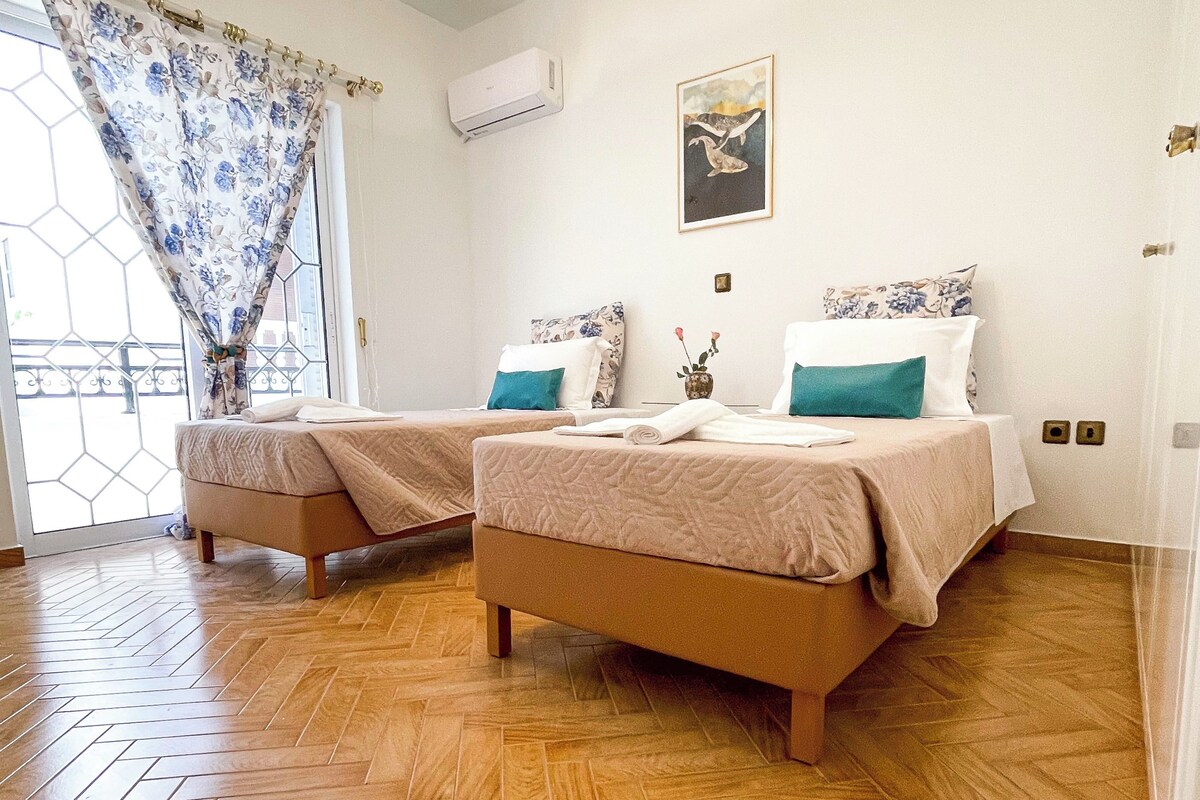 A hotel room with two beds, a ceiling fan, a window with a floral curtain, and a wall-mounted air conditioner.