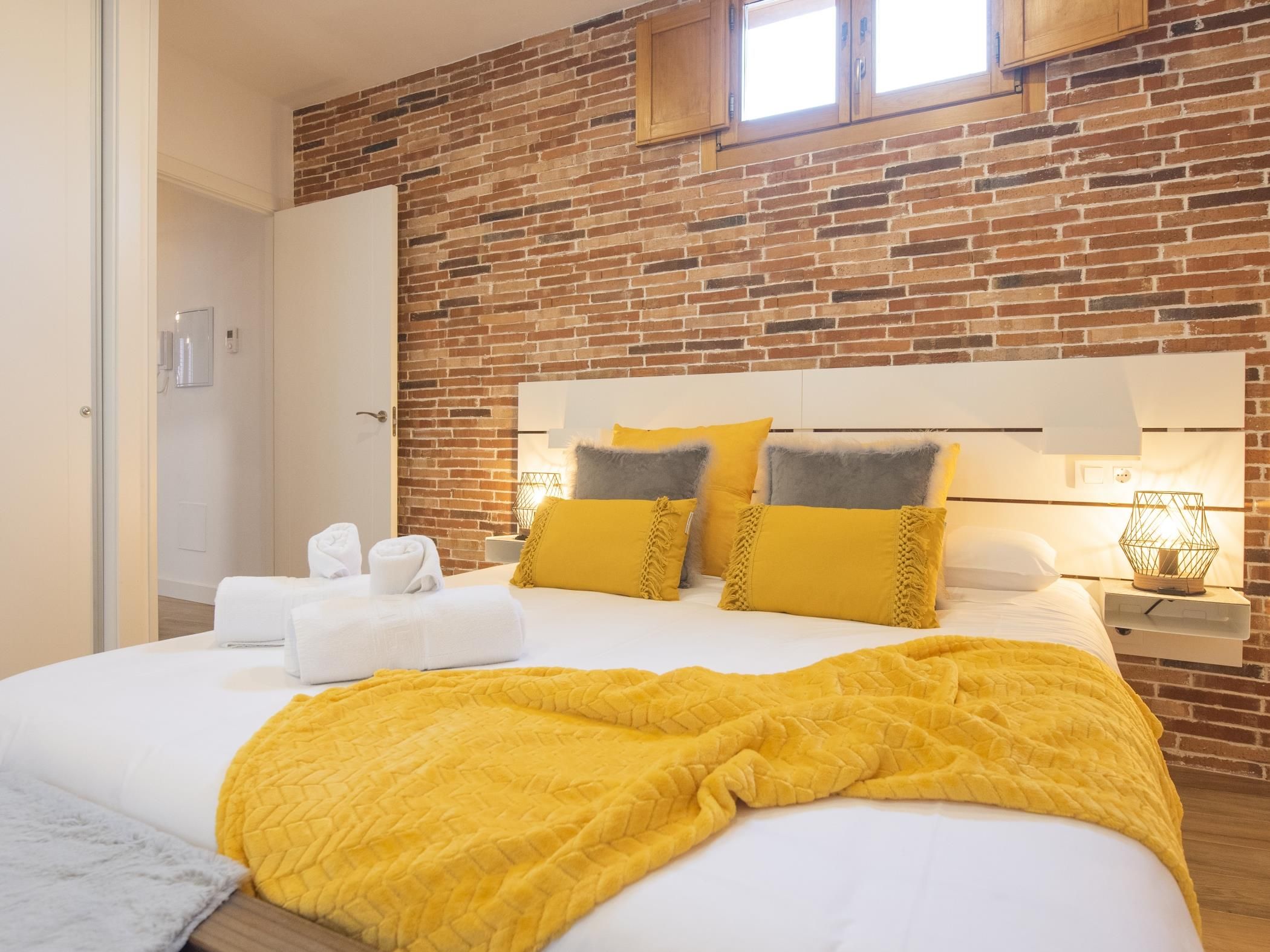 Una habitación de hotel con pared de ladrillo, cama con ropa blanca y manta amarilla.