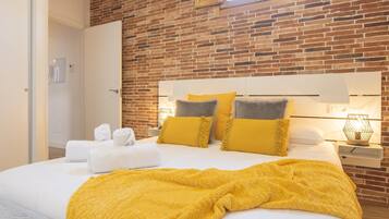 Una habitación de hotel con pared de ladrillo, cama con ropa blanca y manta amarilla.