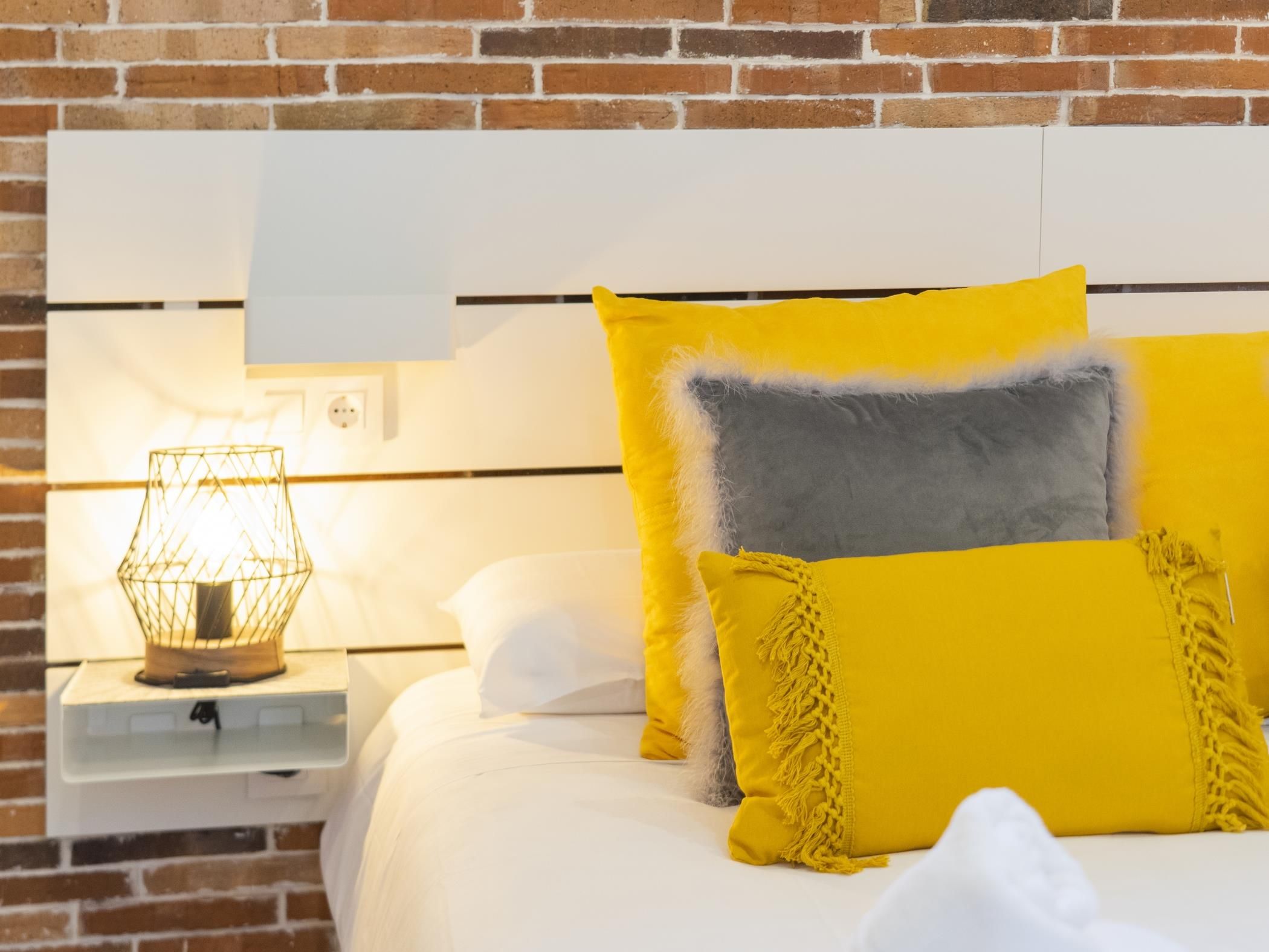 Una cama con ropa blanca y varias almohadas, incluyendo una amarilla y otra gris.