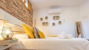 Un dormitorio con pared de ladrillo, una cama con ropa de cama blanca, una mesita de noche con lámpara y un aire acondicionado montado en la pared.