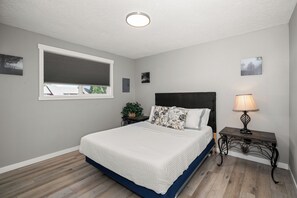 Un dormitorio con cama, mesita de noche, lámpara, ventana con persianas e imágenes colgadas en la pared.