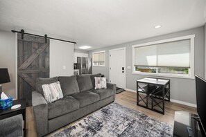 A modern living room with a grey sofa, a coffee table, a TV, and a large window with blinds. - Updated Upstairs 2 Bedroom Apartment - 2 min to Browns Park (Spokane Valley)