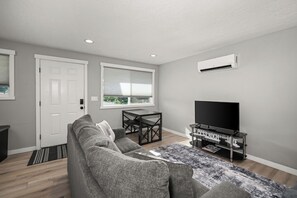 A modern living room with a grey sofa, a coffee table, a TV stand with a flat-screen TV, and an air conditioning unit on the wall.