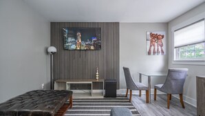 A modern living room with a TV mounted on a slatted wall, a bench, two grey chairs, a small table, and a radiator. - Top Level- Atlanta MBS Apartment w/ Private Hot tub (Atlanta)