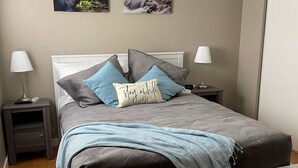 Een slaapkamer met een bed, twee nachtkastjes, een lamp op elk kastje, een deken en twee aan de muur hangende foto's.