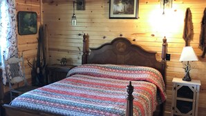 Une chambre chaleureuse dans un chalet en rondins, avec un lit en bois, une lampe rustique, un tableau encadré et une tête de cerf accrochée au mur.