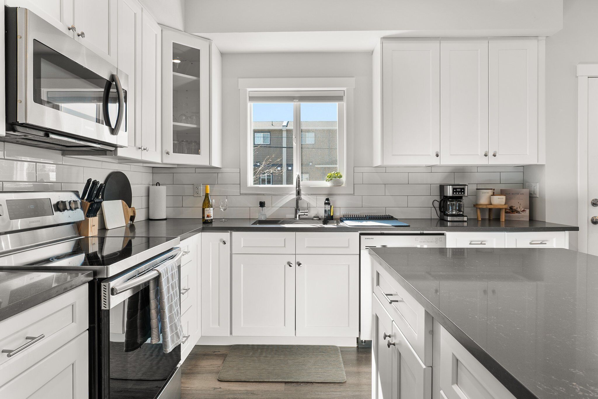 A modern kitchen with white cabinets, stainless steel appliances, a large window, and a black countertop.