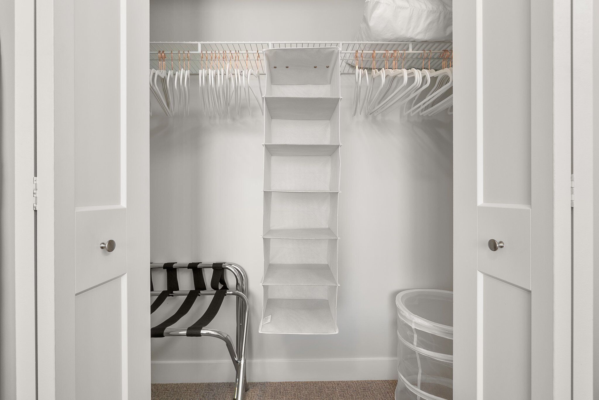 A walk-in closet with open doors, featuring a metal chair, a white shelving unit, and a storage bin.