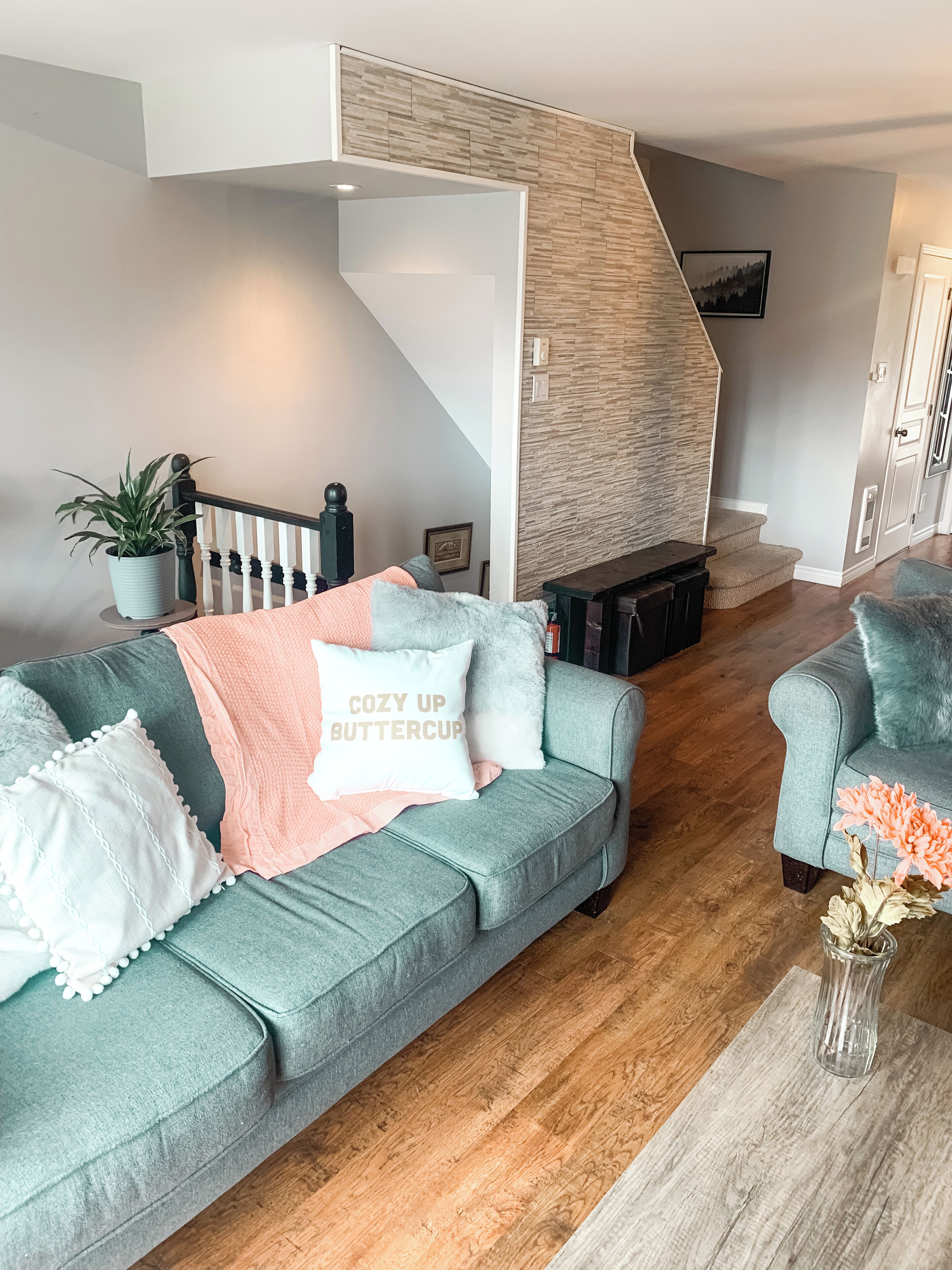 A modern living room with a grey sofa, a coffee table with flowers, a staircase, and a stone wall.