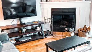 A living room with a flat-screen TV, a fireplace, a sofa, a coffee table, and a bookshelf. - Golf Course Townhome with Deck, BBQ & Views – 4BR Mont Tremblant (Mont-Tremblant)
