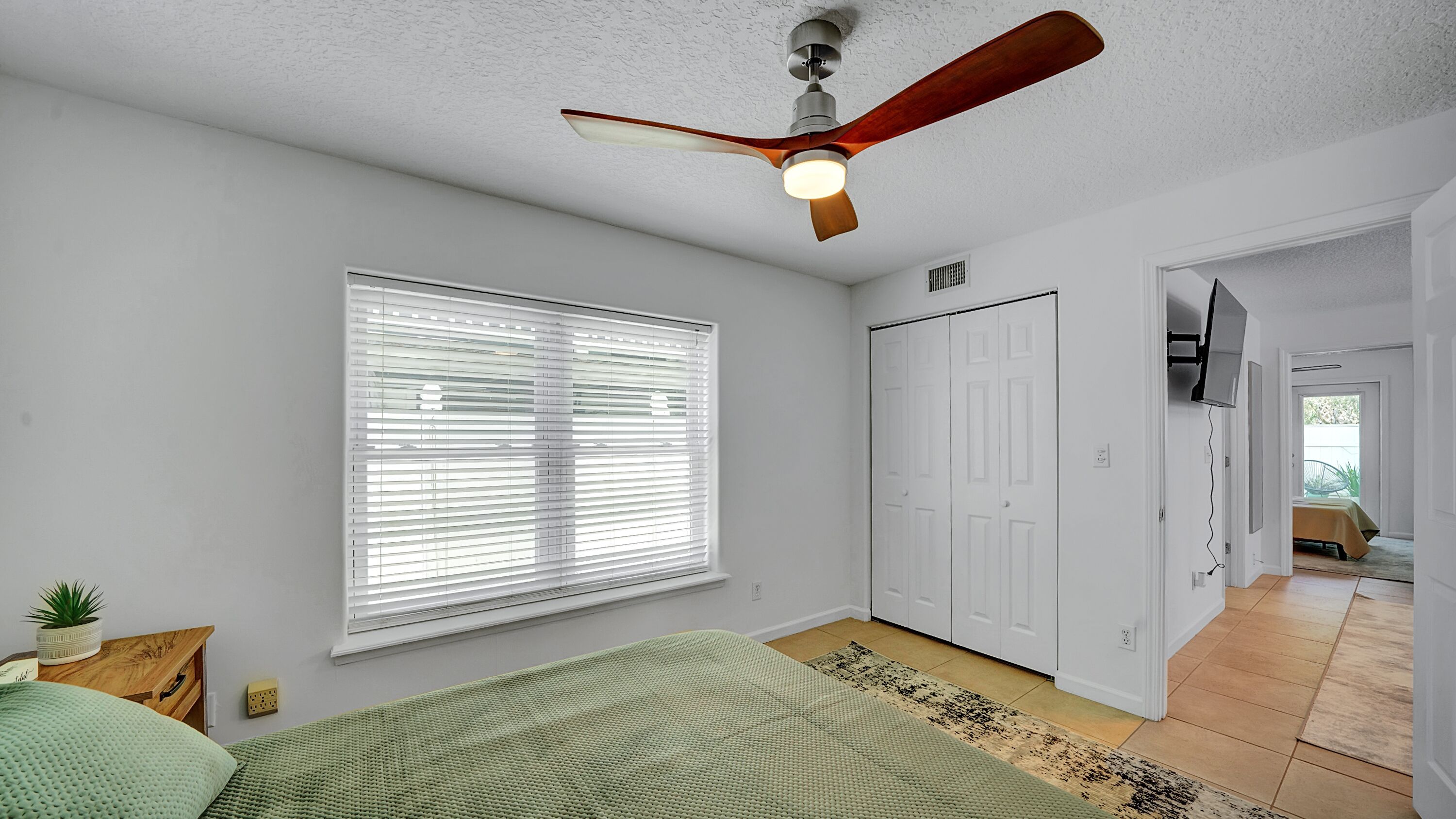 A bedroom with a ceiling fan, a large window with blinds, a bed with green bedding, a white wardrobe, and a TV mounted on the wall.