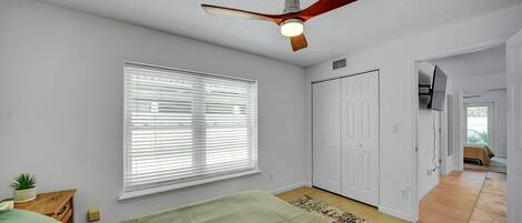 A bedroom with a ceiling fan, a large window with blinds, a bed with green bedding, a white wardrobe, and a TV mounted on the wall.