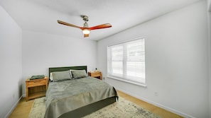 A bedroom with a bed, wooden nightstands, a ceiling fan, a window with blinds, and a patterned rug. - Oceanside 1st floor home, 1 block away from beach and 15 mins away from Mayo (Jacksonville Beach)