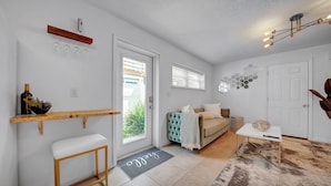 Uma sala de estar moderna com um sofá, uma mesa de centro e uma pequena mesa lateral. Há uma porta que dá para o exterior, uma janela com persianas e um aplique na parede.