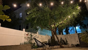 Uma área de pátio ao ar livre com luzes de corda, uma árvore e um cercado branco.