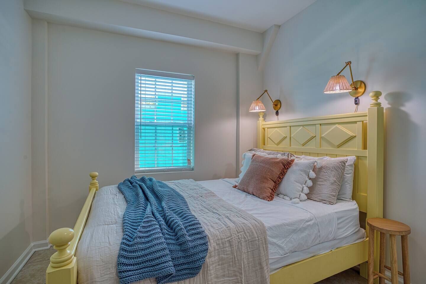 Un dormitorio con una cama, una ventana con persianas, dos lámparas de pared y un taburete de madera.