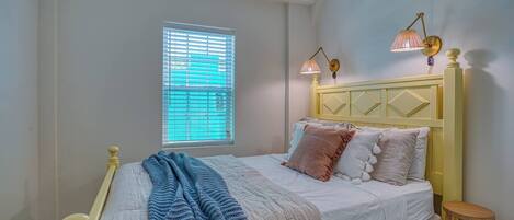 Un dormitorio con una cama, una ventana con persianas, dos lámparas de pared y un taburete de madera.