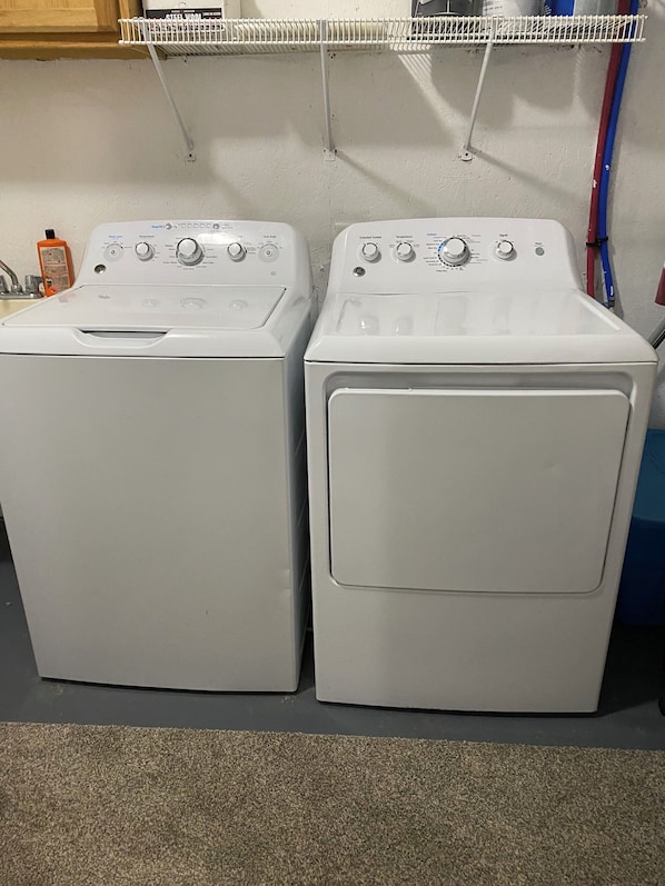 Two front-loading white washing machines in a laundry room with a metal shelf above and a bottle of detergent on the left.