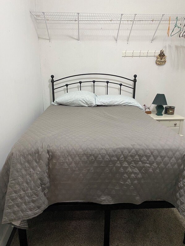 Un letto singolo con copripiumino grigio trapuntato, una testiera in metallo nero e un comodino bianco con una lampada verde e una piccola statuina.