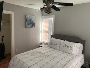 A bedroom with a bed, ceiling fan, artwork, and a window with blinds.