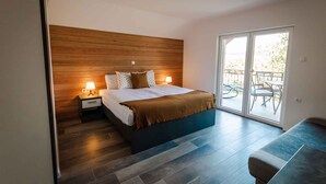 Un dormitorio con cabecera de madera, cama con ropa de cama blanca, mesita de noche con lámpara y balcón con mobiliario exterior.