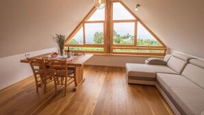 Een eethoek met een houten tafel en stoelen, een bank en grote ramen met uitzicht op groen.