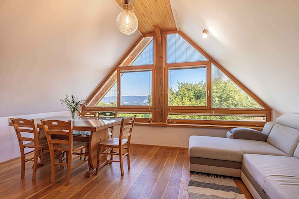 A dining area with a wooden table and chairs, a sofa, and large windows with a view of greenery.