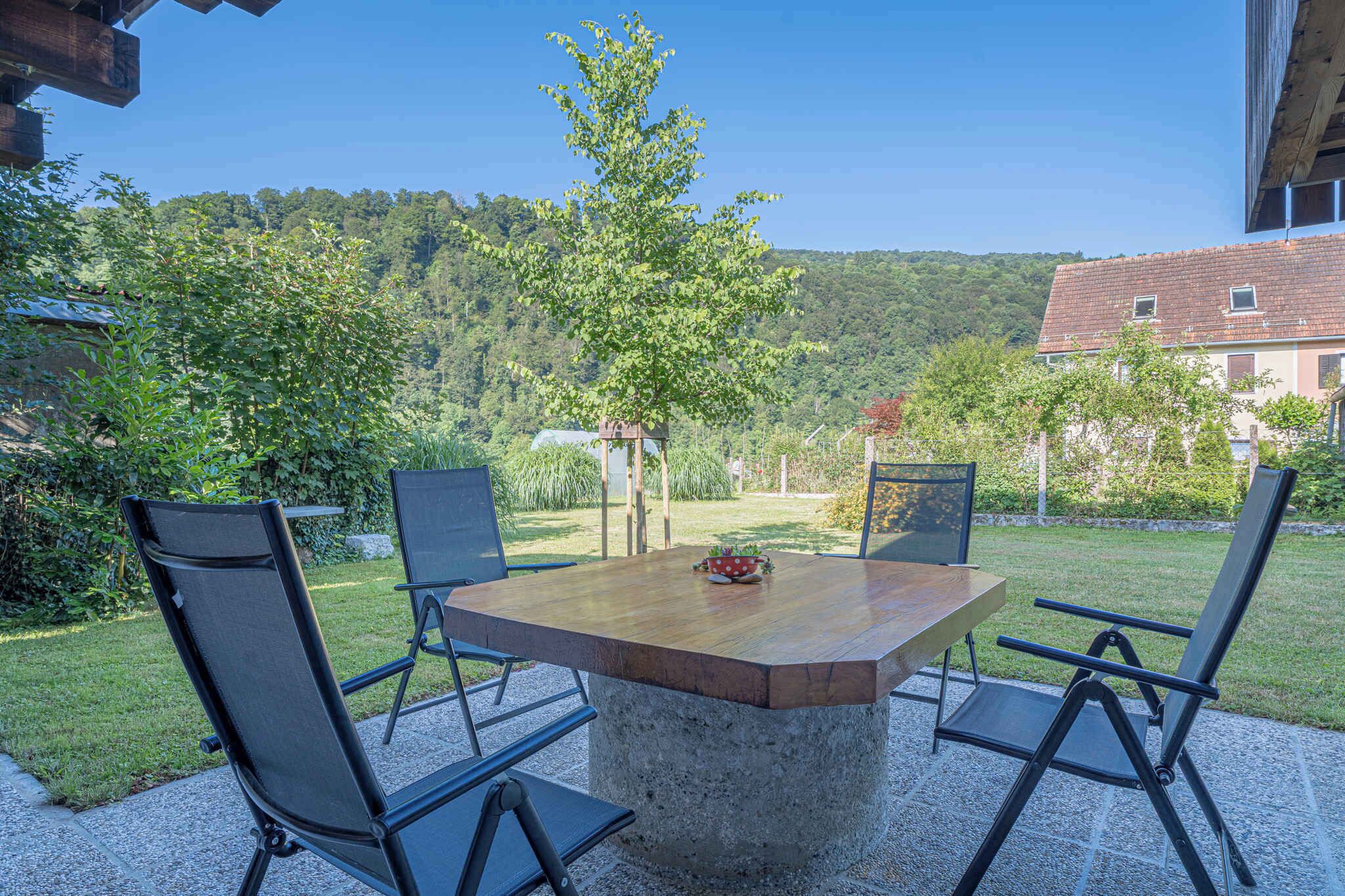 An outdoor patio with a concrete table and four chairs, surrounded by a lush garden and a view of a forested hill.