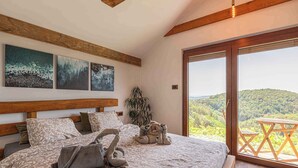 Une chambre à coucher avec un grand lit, une tête de lit en bois et une vue sur une colline boisée, visible à travers une porte-fenêtre coulissante.
