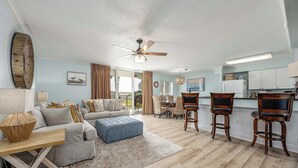 A spacious living room with a sofa, ottoman, and wooden chairs, a dining area with a table and chairs, and a kitchen with a refrigerator and bar stools.