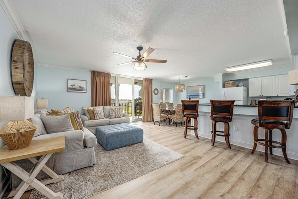 A spacious living room with a sofa, ottoman, and wooden chairs, a dining area with a table and chairs, and a kitchen with a refrigerator and bar stools.