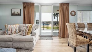 A living room with a sofa, patterned cushions, a dining table with chairs, and a view of a pool and palm trees through a sliding glass door.