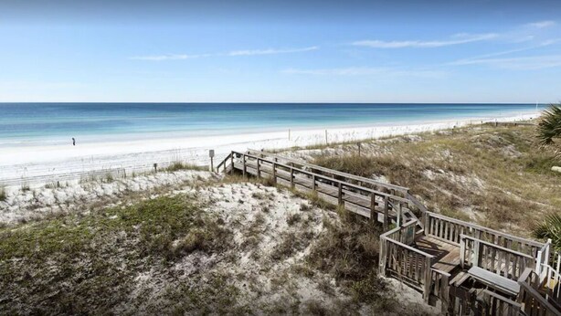 A beach with a wooden boardwalk leading to the shore, sandy dunes, and a clear blue sky.