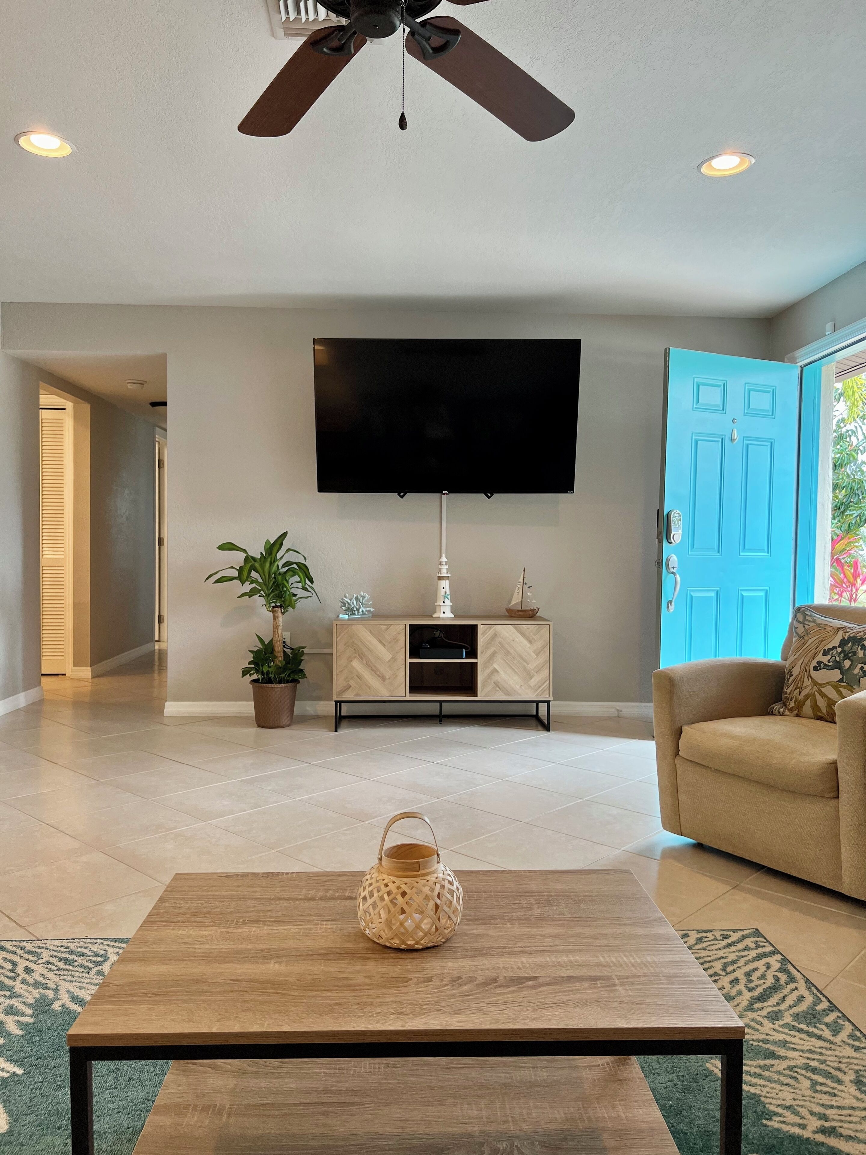 A living room with a ceiling fan, a flat-screen TV, a wooden coffee table, a beige sofa, and a blue door.