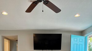 Een woonkamer met een plafondventilator, een flatscreen televisie, een houten salontafel, een beige bank en een blauwe deur.