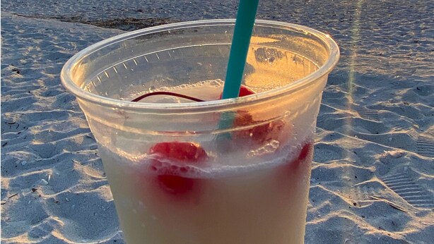 Een plastic beker met rietje en rood fruit op een strand, met de ondergaande zon op de achtergrond.