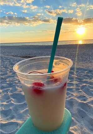Ein Plastikbecher mit Trinkhalm und roten Früchten an einem Strand, im Hintergrund ein Sonnenuntergang.