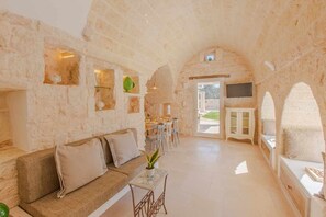 A cozy living area with a sofa, a coffee table, and a TV. There is a dining area with chairs and a table. The room has stone walls and an arched ceiling.