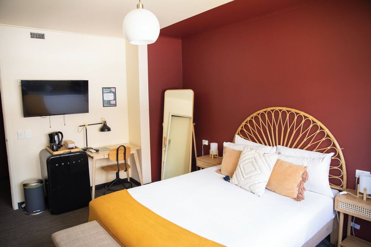 A hotel room with a bed, a desk, a chair, a television, and a trash bin.