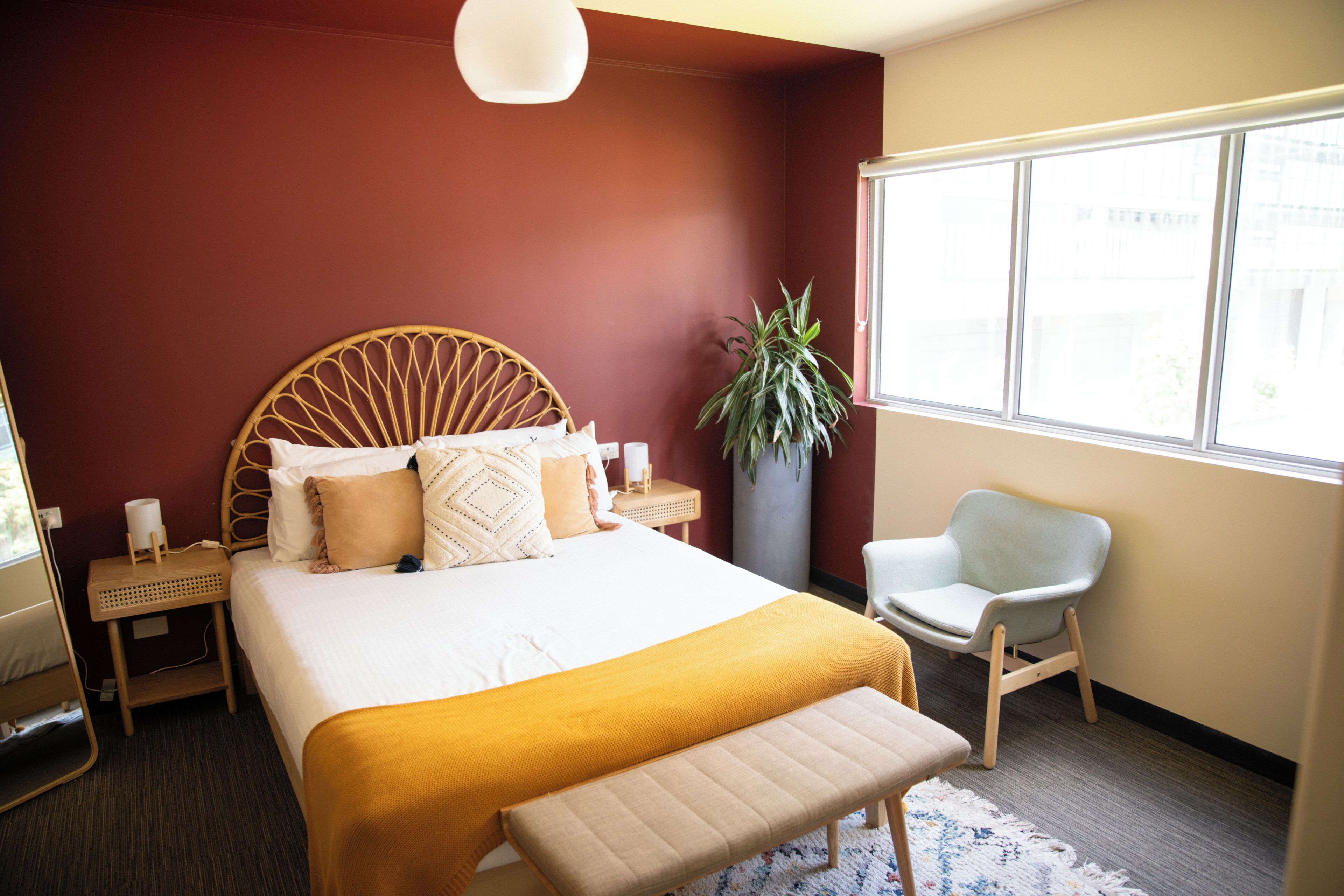 Una habitación de hotel con cama, mesitas de noche, una silla, un ventanal grande y una planta en maceta.