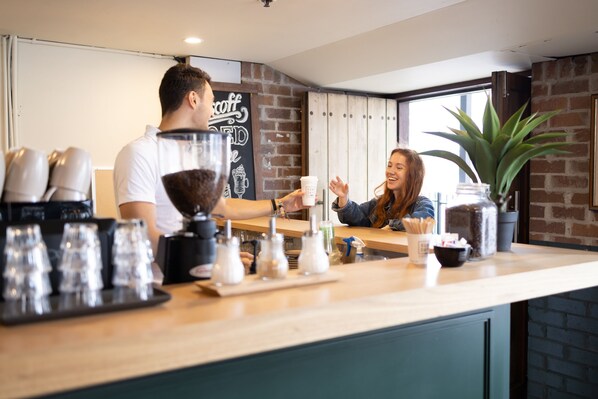 Mostrador de cafetería con un barista y un cliente. Hay tazas de café, un espumador de leche y una planta en el mostrador.