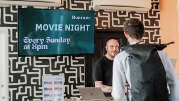 A room with a patterned wall, a television displaying 'Roamer MOVIE NIGHT Every Sunday at 8pm', and a table with a laptop and brochures.