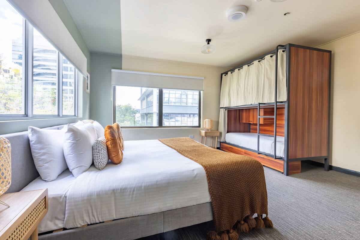 A modern hotel room with a large bed, a bunk bed, and a window with a view of buildings.