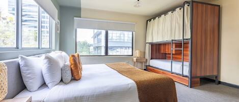 A modern hotel room with a large bed, a bunk bed, and a window with a view of buildings.