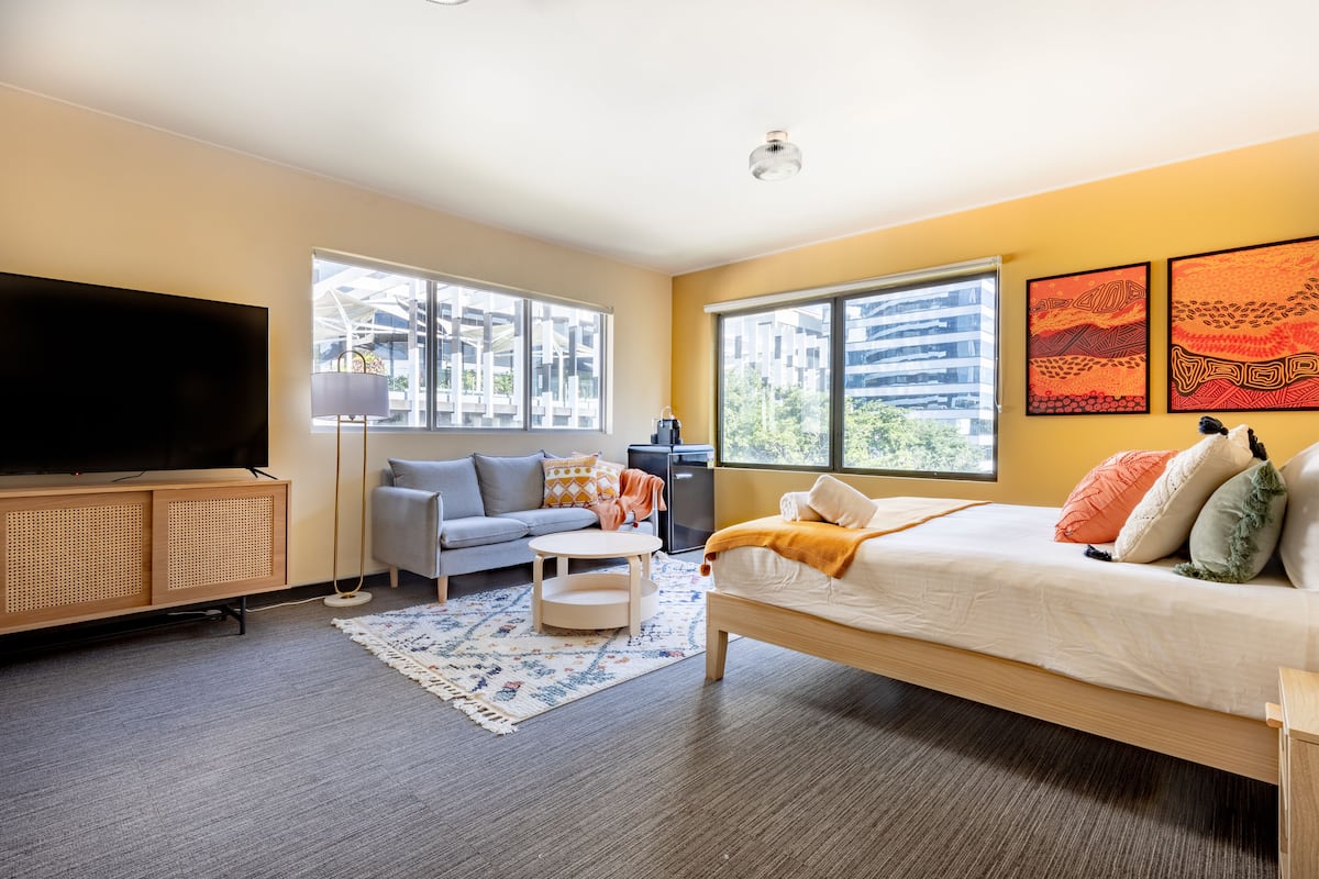 A modern hotel room with a large bed, a sofa, a TV, and large windows offering a city view.