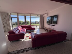 A modern living room with a red sectional sofa, a flat-screen TV mounted on the wall, and large windows offering a view of the outdoors. - The Cutting Estate Mount Panorama (Mount Panorama)