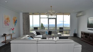 A modern living room with a sectional sofa, a chandelier, large windows with a view, and artwork on the walls. - The Cutting Estate Mount Panorama (Mount Panorama)