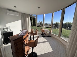 A room with a kitchen area, a dining table with chairs, and large windows offering a view of a garden.